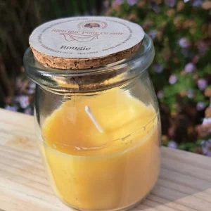 Bougie jaune dans son petit pot en verre avec un couvercle en liège, sur une planche de bois, fond jardin fleuris
