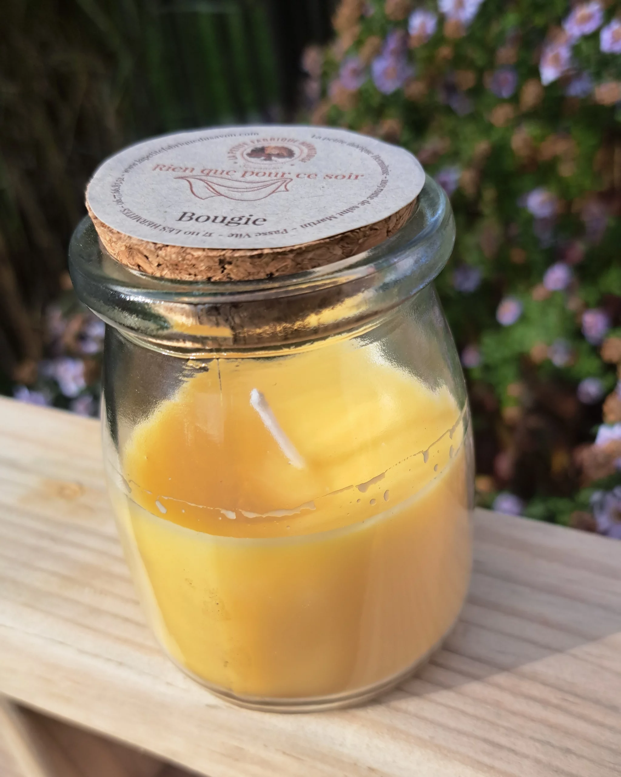 Bougie jaune dans son petit pot en verre avec un couvercle en liège, sur une planche de bois, fond jardin fleuris